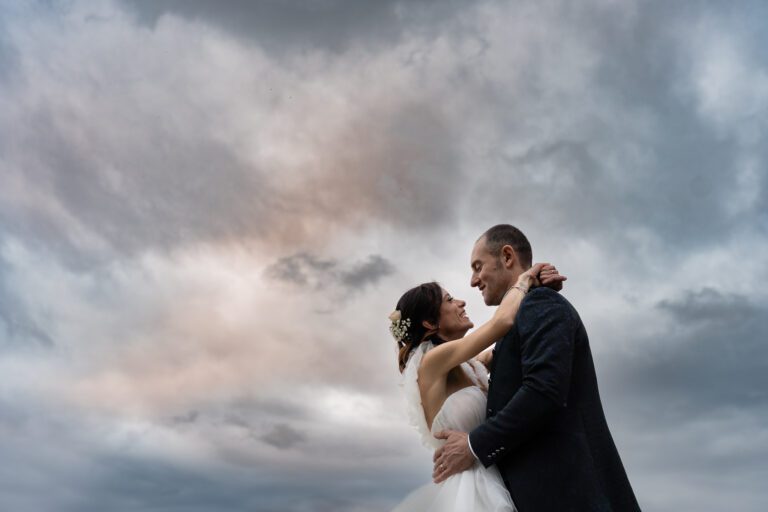 Gli sposi si abbracciano sotto un drammatico cielo nuvoloso, condividendo un momento d'amore nel loro foto matrimonio. La sposa indossa un abito bianco, lo sposo un abito scuro.