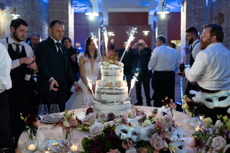 Una coppia di sposini guarda una torta nuziale a tre livelli adornata con fiori e stelle filanti, con gli ospiti che guardano in un luogo rustico poco illuminato.