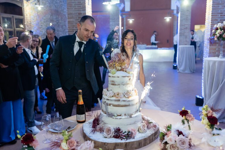 Una sposa e uno sposo ridono gioiosamente mentre tagliano una torta nuziale a più livelli durante il loro ricevimento di matrimonio, circondati da ospiti applaudinti in una sala addobbata a festa.