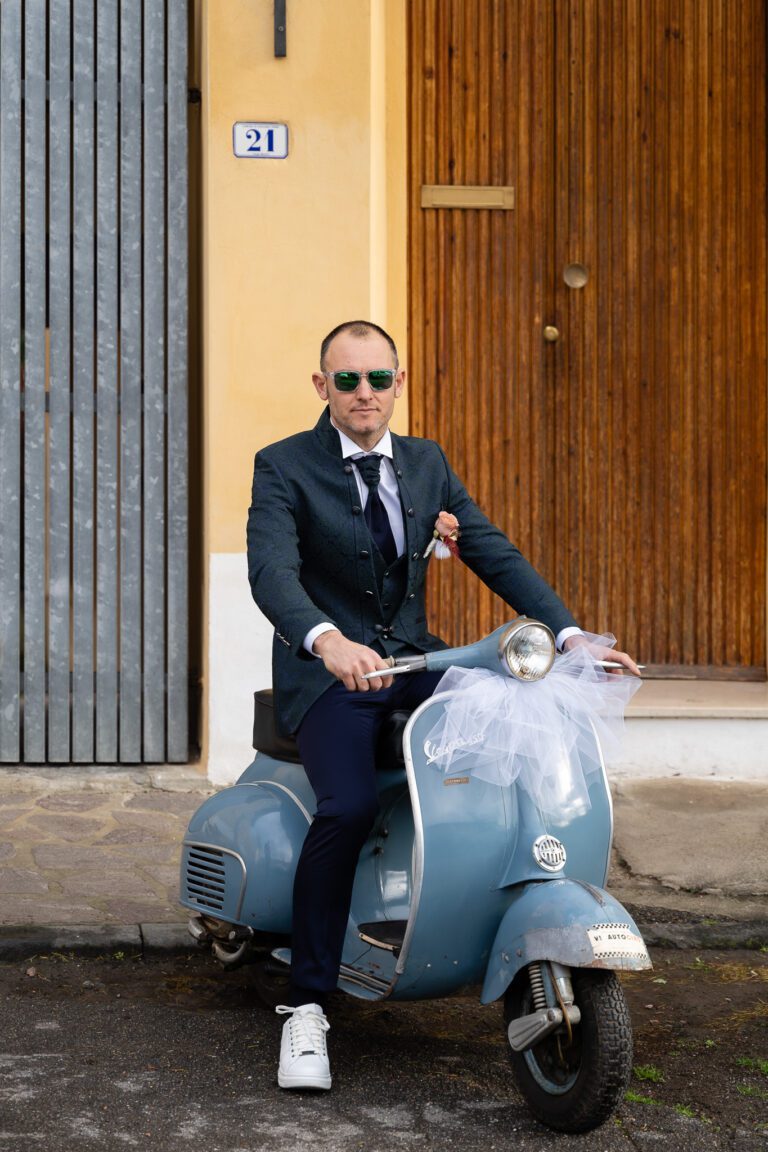 Un uomo ben vestito in abito e occhiali da sole si siede su uno scooter blu vintage davanti a un muro giallo con porte per una foto di matrimonio.