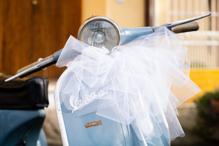 Foto di matrimonio in primo piano di uno scooter Vespa azzurro con un grande fiocco di tulle bianco sul manubrio, che evidenzia il marchio "Vespa" e il nome del modello "Primavera"