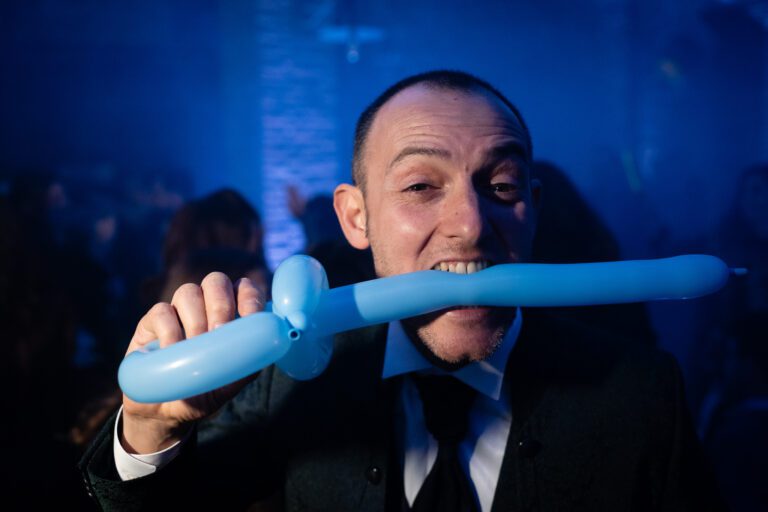 Un uomo a una festa morde un palloncino azzurro attorcigliato, facendo una faccia giocosa. la scena è vivace con uno sfondo affollato illuminato di blu.