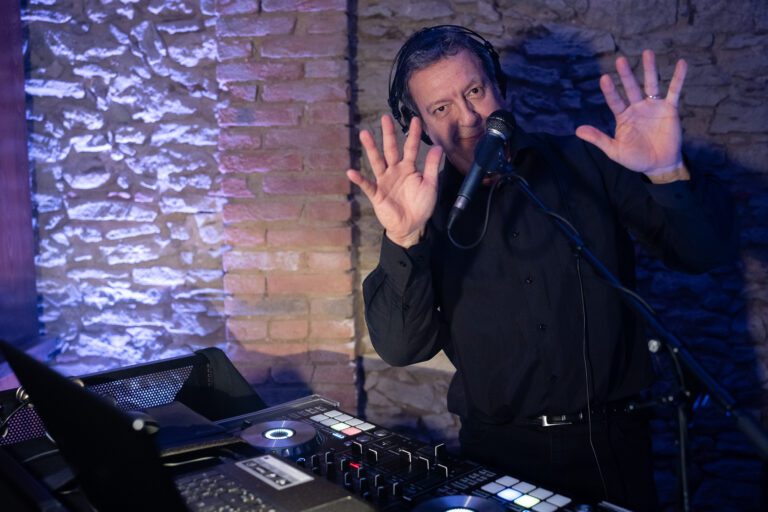 Un dj maschio di mezza età con una camicia nera che gesticola mentre mixa musica su una console in una stanza scarsamente illuminata con pareti di mattoni.