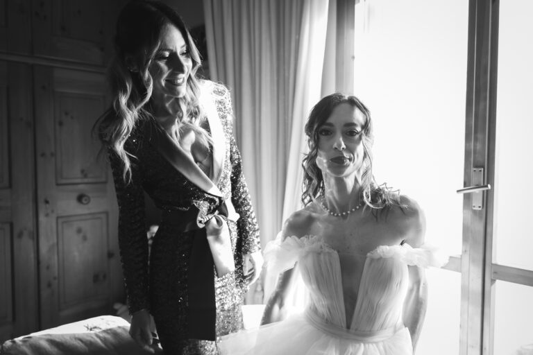 Foto di matrimonio in bianco e nero di due donne sorridenti, una con un glamour abito di paillettes e l'altra con un elegante abito da sposa, in piedi all'interno vicino a una grande finestra.