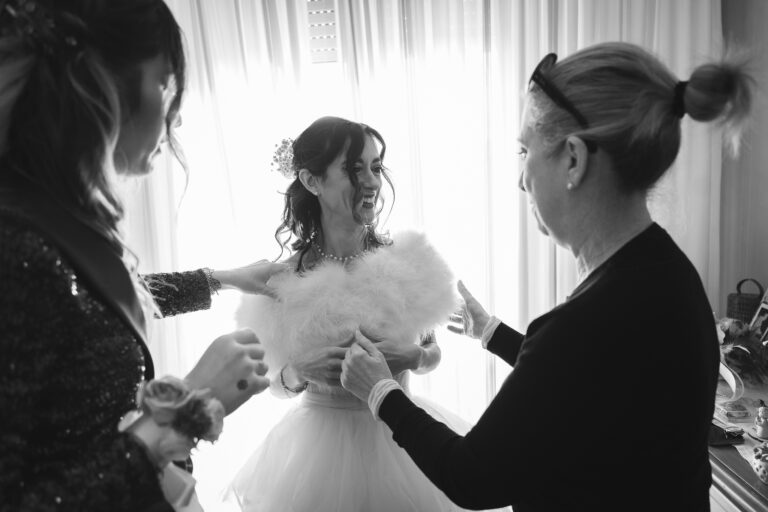 Una sposa in abito bianco sorride gioiosamente mentre viene aiutata da due donne, probabilmente parenti strette o amiche, che le stanno aggiustando il vestito in una stanza ben illuminata per una fotografia.