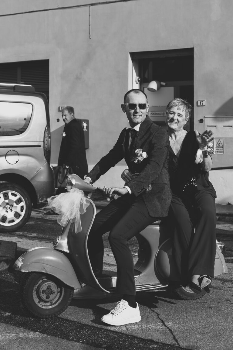 Una coppia gioiosa, vestita elegantemente con bottoniere, guida una vespa bianca dopo il matrimonio, sorridendo ampiamente sotto la luce del sole nella loro foto di matrimonio, con gli edifici sullo sfondo.