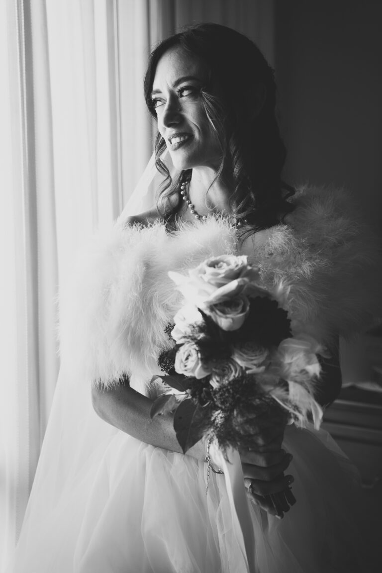 Una sposa radiosa in abito bianco, con in mano un bouquet e avvolta in una stola di piume, sorride con gioia mentre guarda fuori da una finestra. La foto matrimonio è in bianco e nero