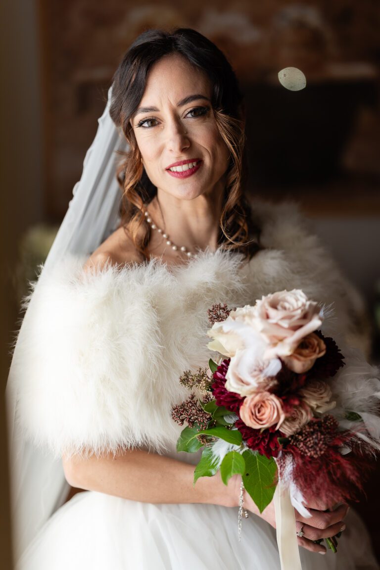 Una sposa in abito bianco e stola di pelliccia tiene in mano un mazzo di rose, sorride dolcemente all'interno con luci soffuse che mettono in risalto i suoi lineamenti in questo incantevole foto di matrimonio.