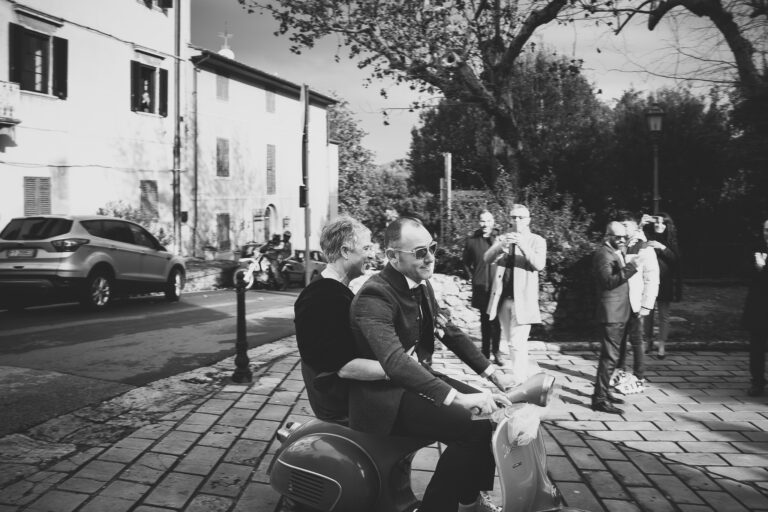 Due uomini in giacca e cravatta guidano uno scooter vintage su una strada soleggiata fiancheggiata da alberi ed edifici, sorridendo mentre gli spettatori osservano e catturano foto di questo gioioso momento di matrimonio.