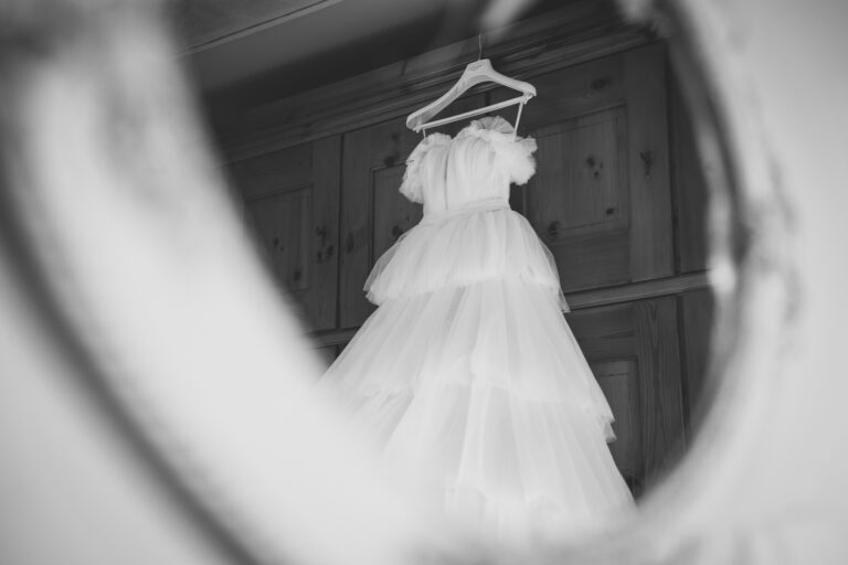 Una foto in bianco e nero di un abito da sposa appeso all'anta di un armadio, visto attraverso la cornice ovale di uno specchio, creando un'atmosfera artistica e vintage.